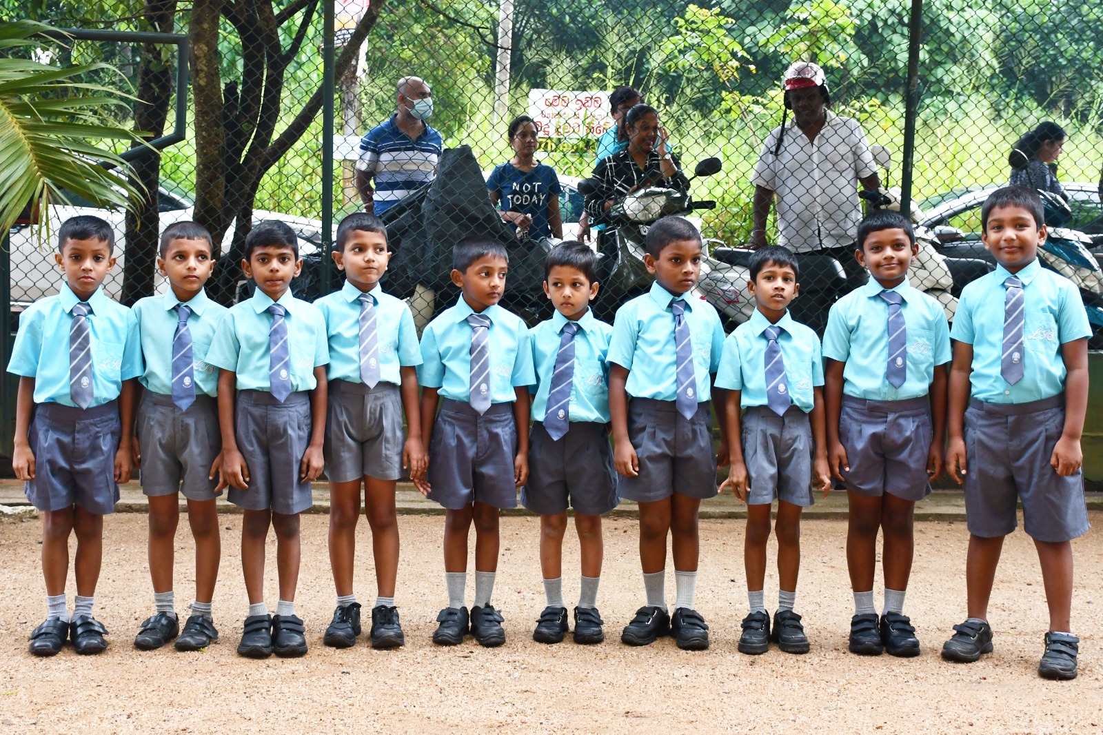 Young schoolboys in uniform standing outdoors with adults in the background.