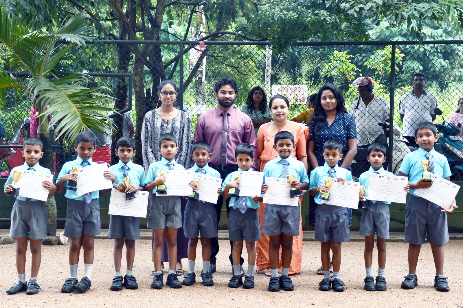 Young students in blue uniforms with awards, teachers behind, green playground setting.