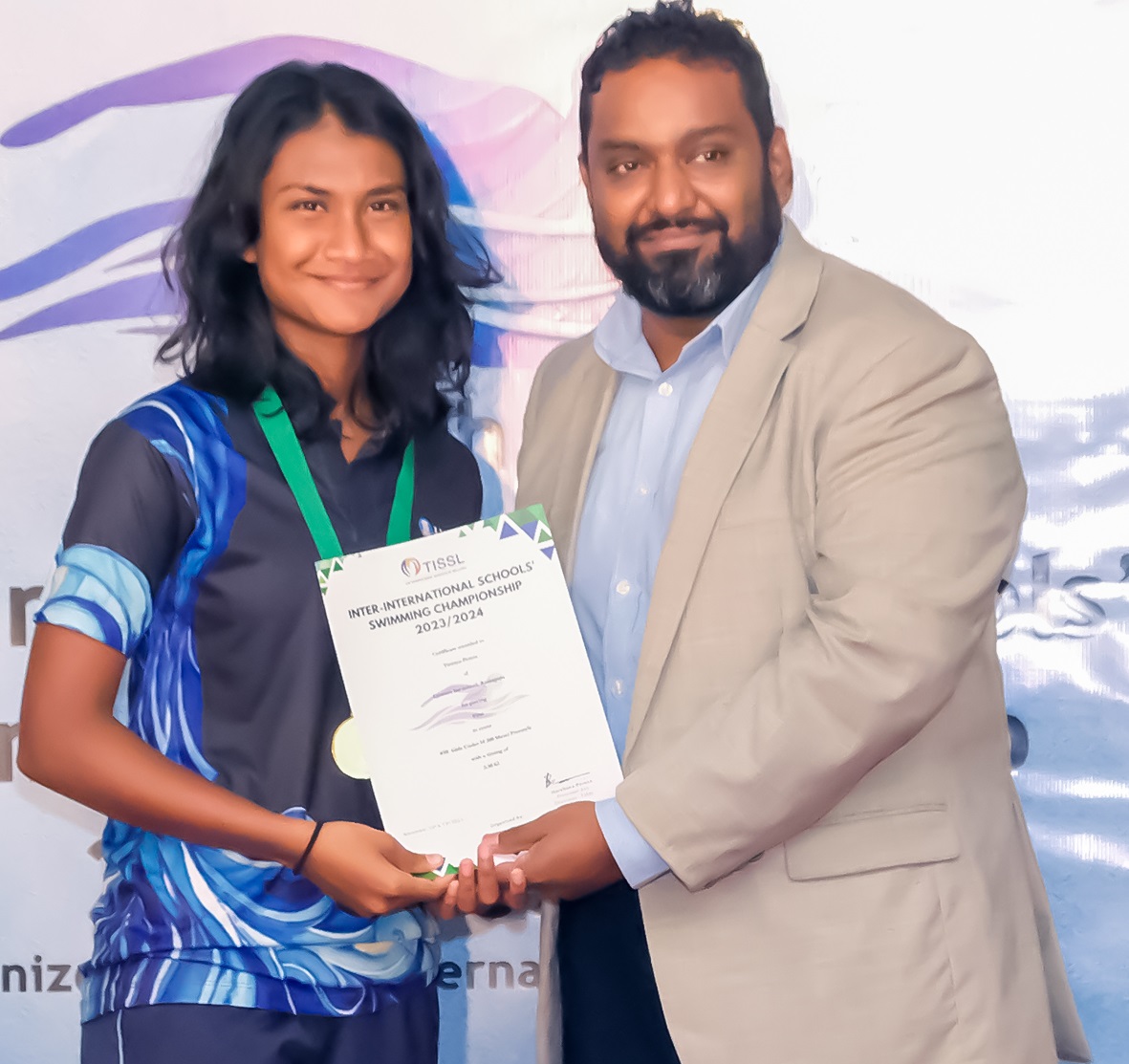 Young woman receives award at swimming championship ceremony, presented by bearded man in light blazer.