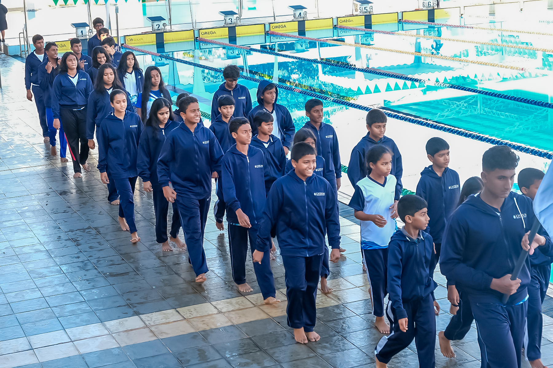 Swim team in blue tracksuits walking beside indoor pool, preparing for practice.