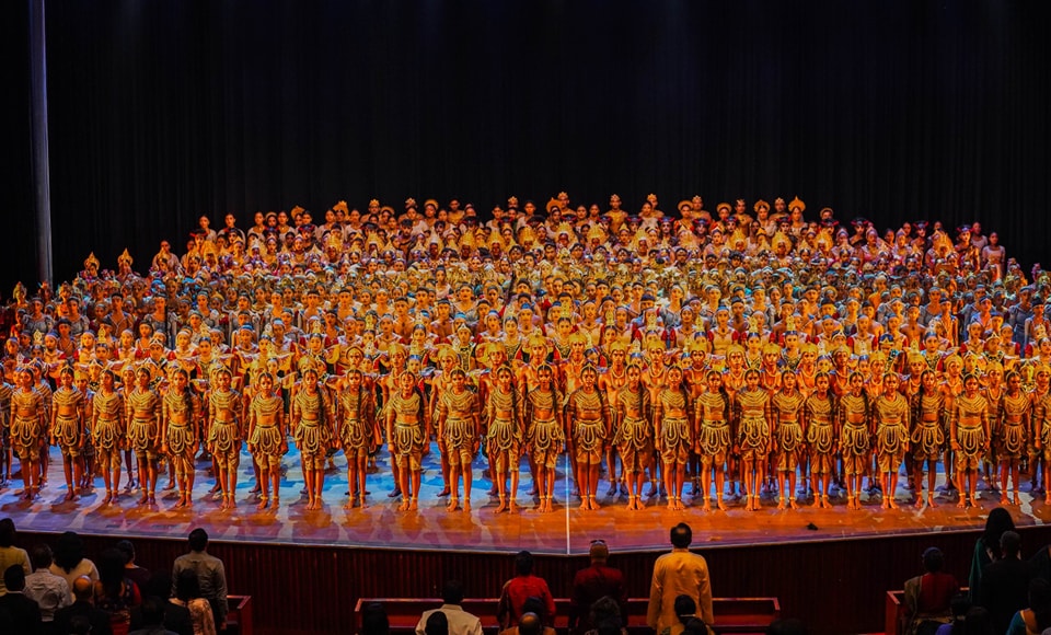 Grand stage with performers in vibrant traditional costumes, bright lighting, reflective surface, dark backdrop.