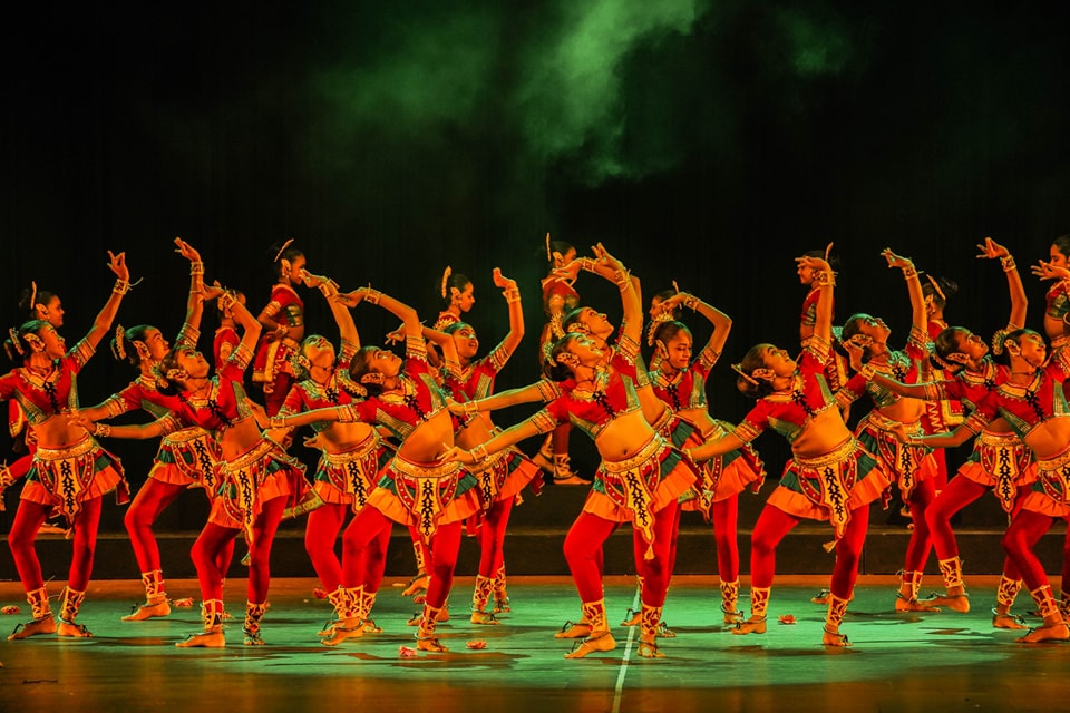 Synchronized dancers in vibrant costumes perform on a dramatically lit stage with green fog.