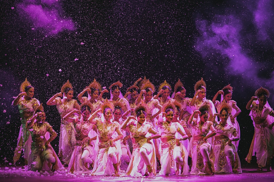 Dancers in traditional attire perform against a purple, smoky backdrop, creating a mystical ambiance.