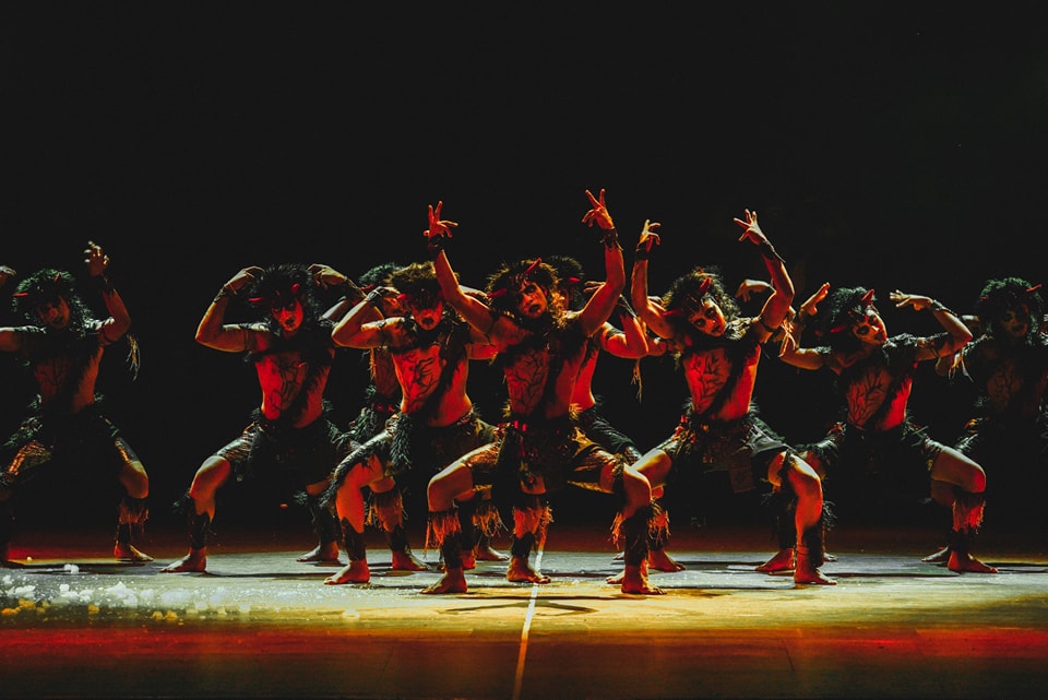Tribal dancers in powerful poses on a dimly lit stage with dramatic reddish lighting.