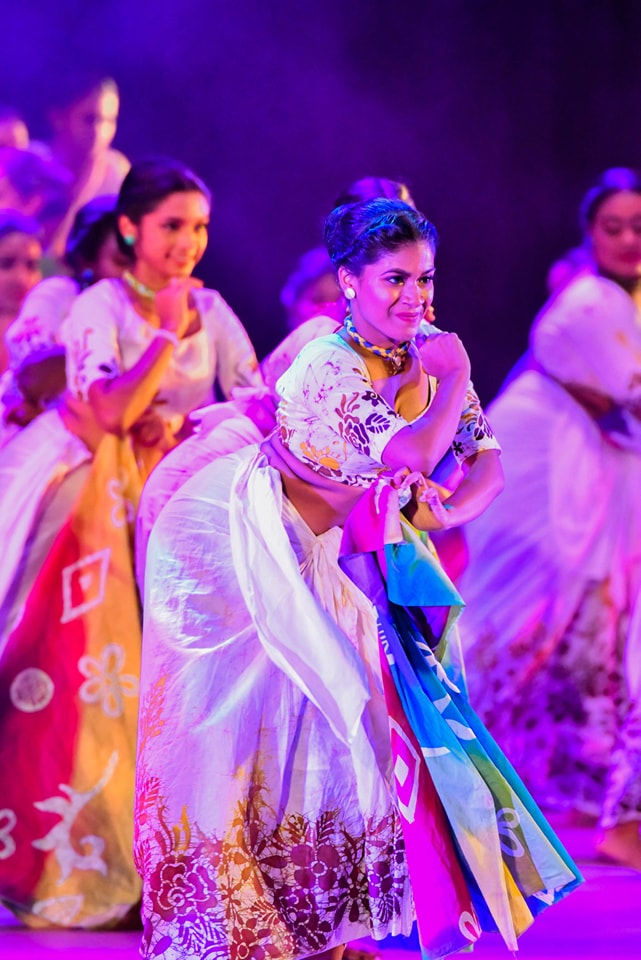Women dancing in colorful traditional attire under vibrant purple stage lighting.