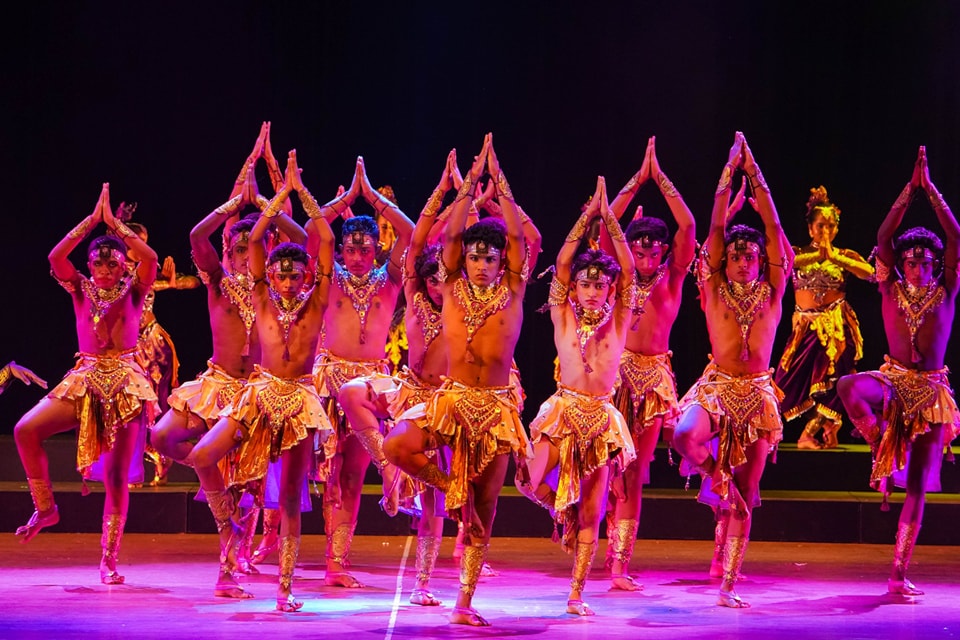 Traditional dancers in gold and yellow costumes perform a synchronized routine on a vibrantly lit stage.