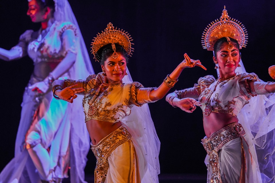 Traditional dancers in ornate gold costumes and headdresses perform expressive dance on stage.