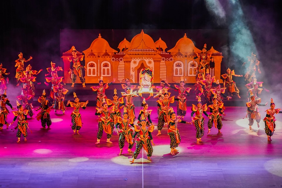 Cultural dancers in colorful costumes perform on a dramatically lit stage with an ornate backdrop.