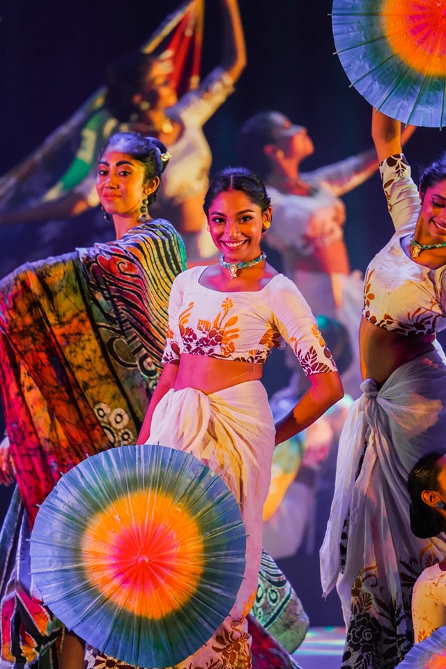 Traditional dancers in colorful attire perform with vibrant umbrellas on a warmly lit stage.