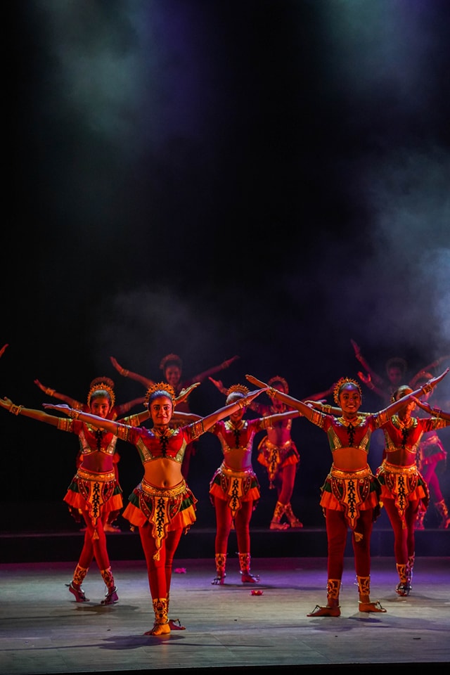 Dancers in vibrant traditional costumes perform on a misty, well-lit stage.