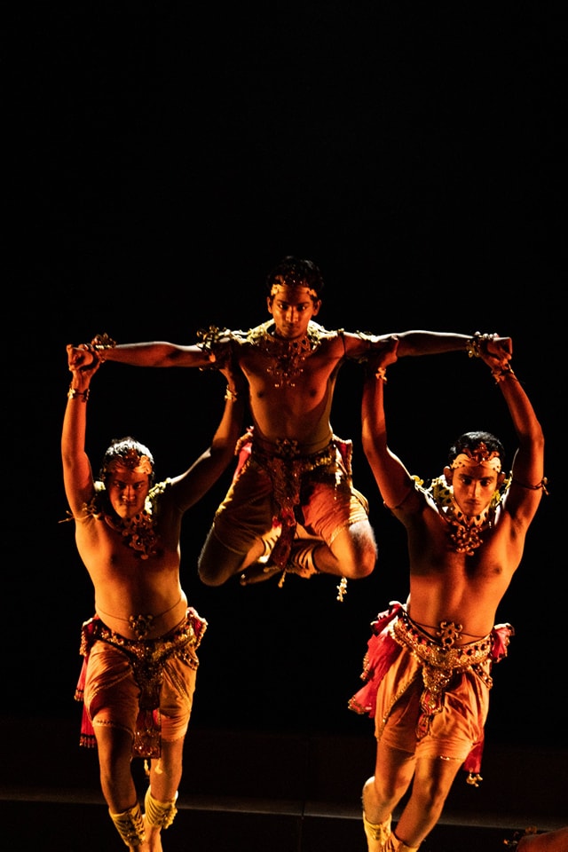 Three male dancers in vibrant traditional attire performing an acrobatic dance against a dark background.