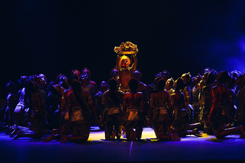 Central figure holds sacred artifact as performers kneel in traditional attire under spotlight.