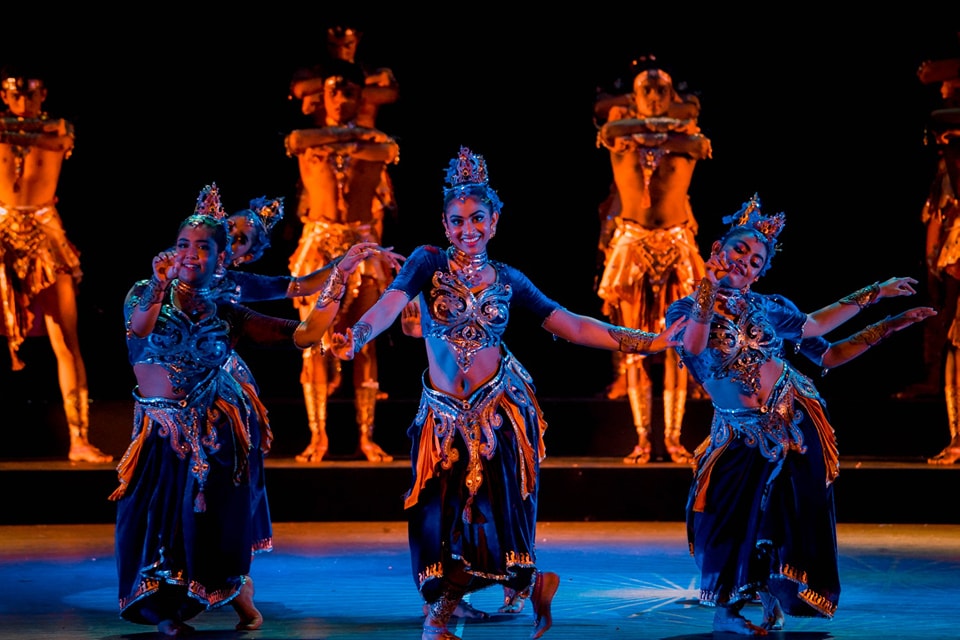 Traditional dancers in ornate blue and gold costumes perform energetically on a dimly lit stage.