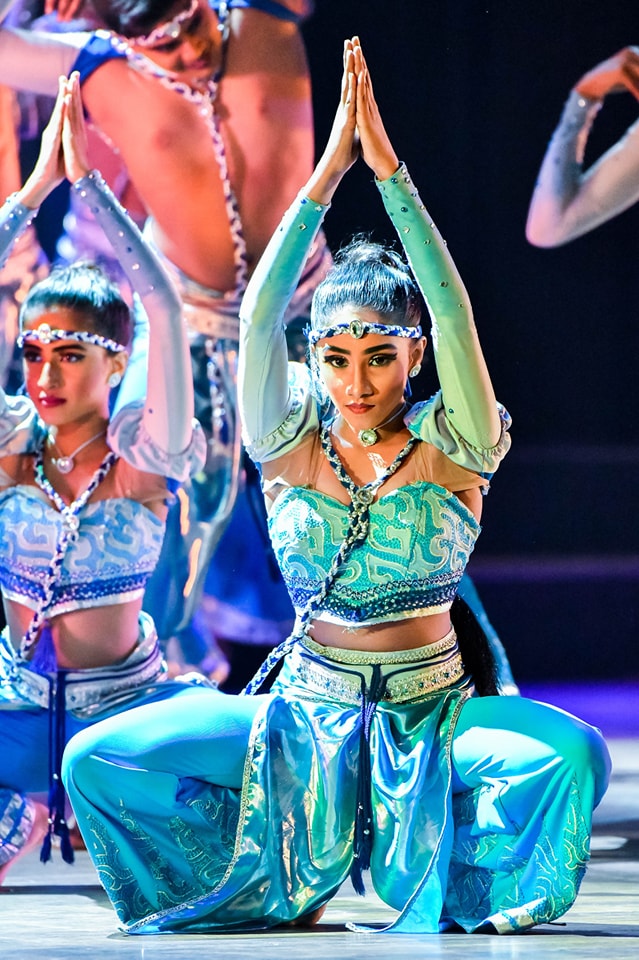 Central dancer in vibrant turquoise costume kneeling, hands in prayer, surrounded by fellow performers.