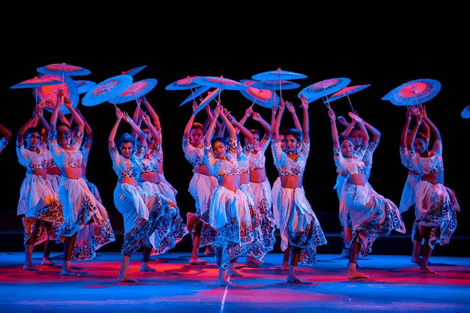 Traditional dancers in colorful costumes with circular props, blue and red stage lighting.