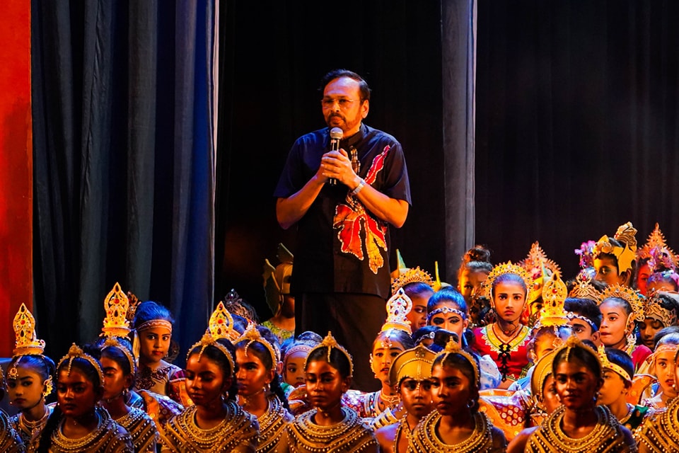 Traditional cultural performance with speaker and young girls in ornate attire and headpieces.