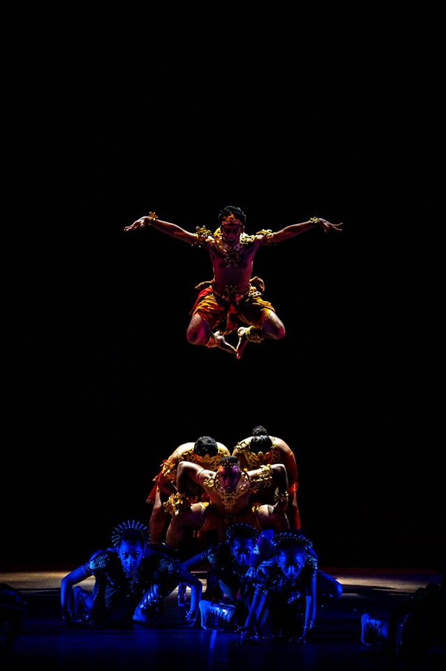 Airborne dancer in vibrant costume above a supportive human pyramid in dramatic lighting.