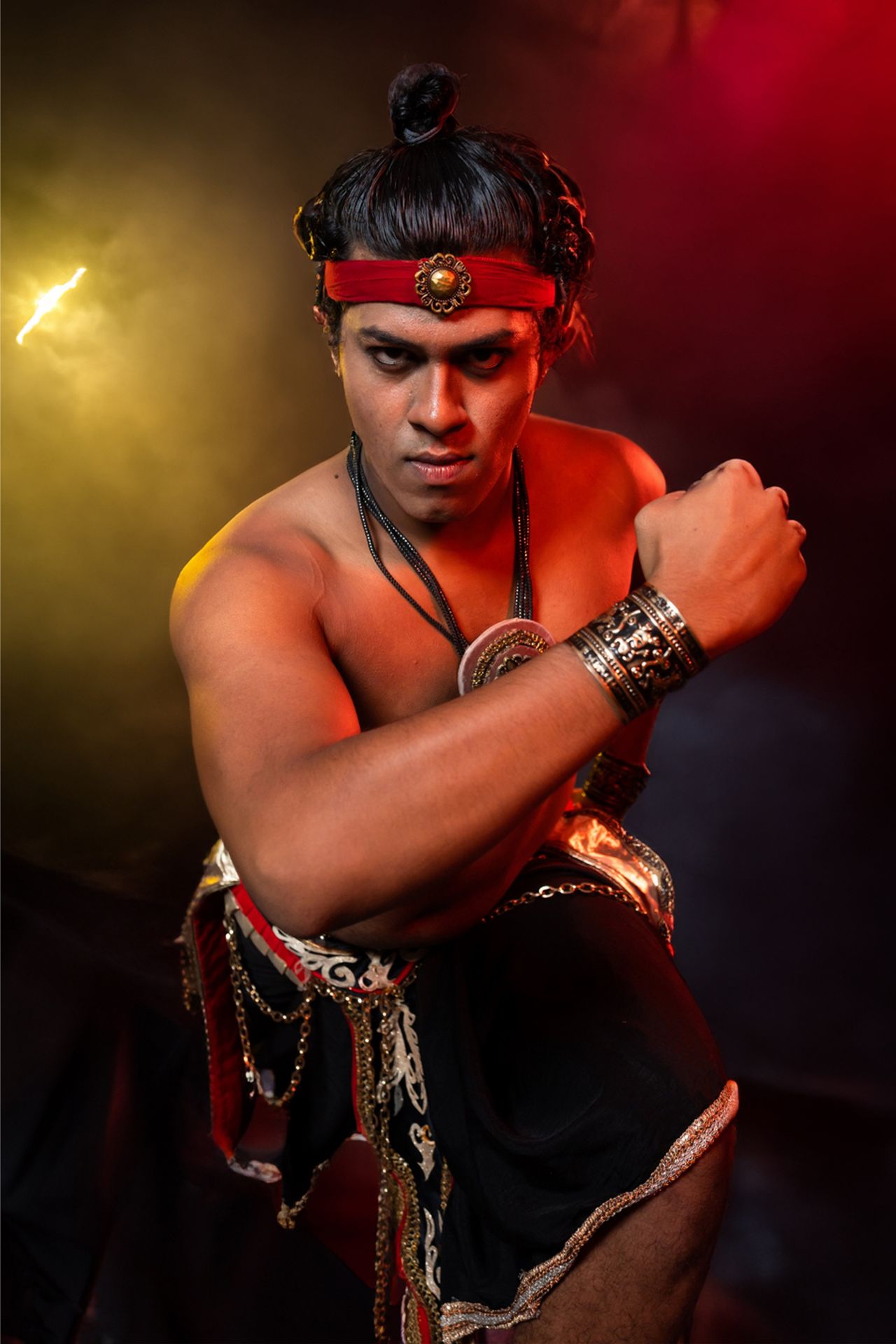 A warrior in traditional attire poses intensely with a smoky red and black background.