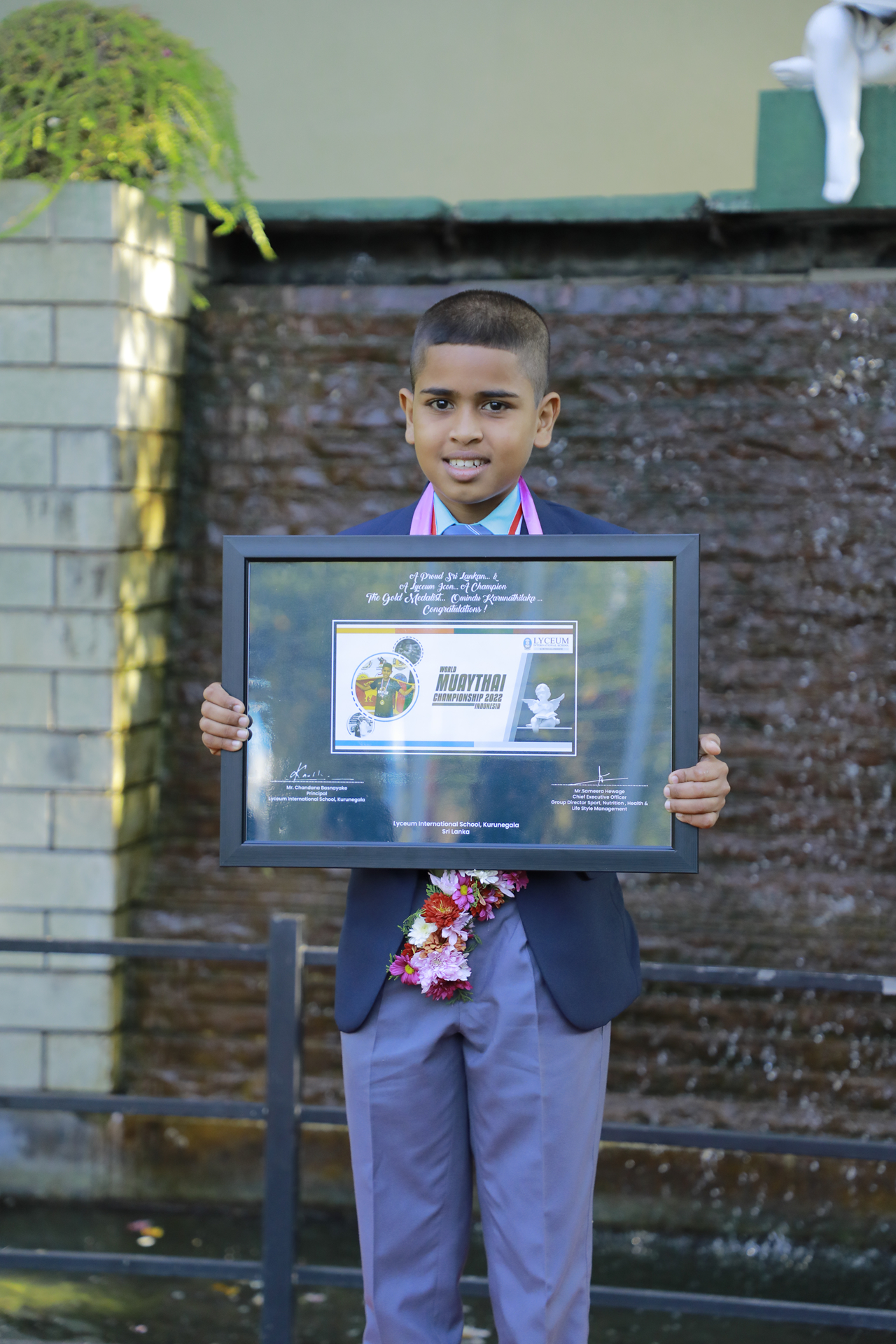 Young boy proudly holds award in front of a serene water feature.