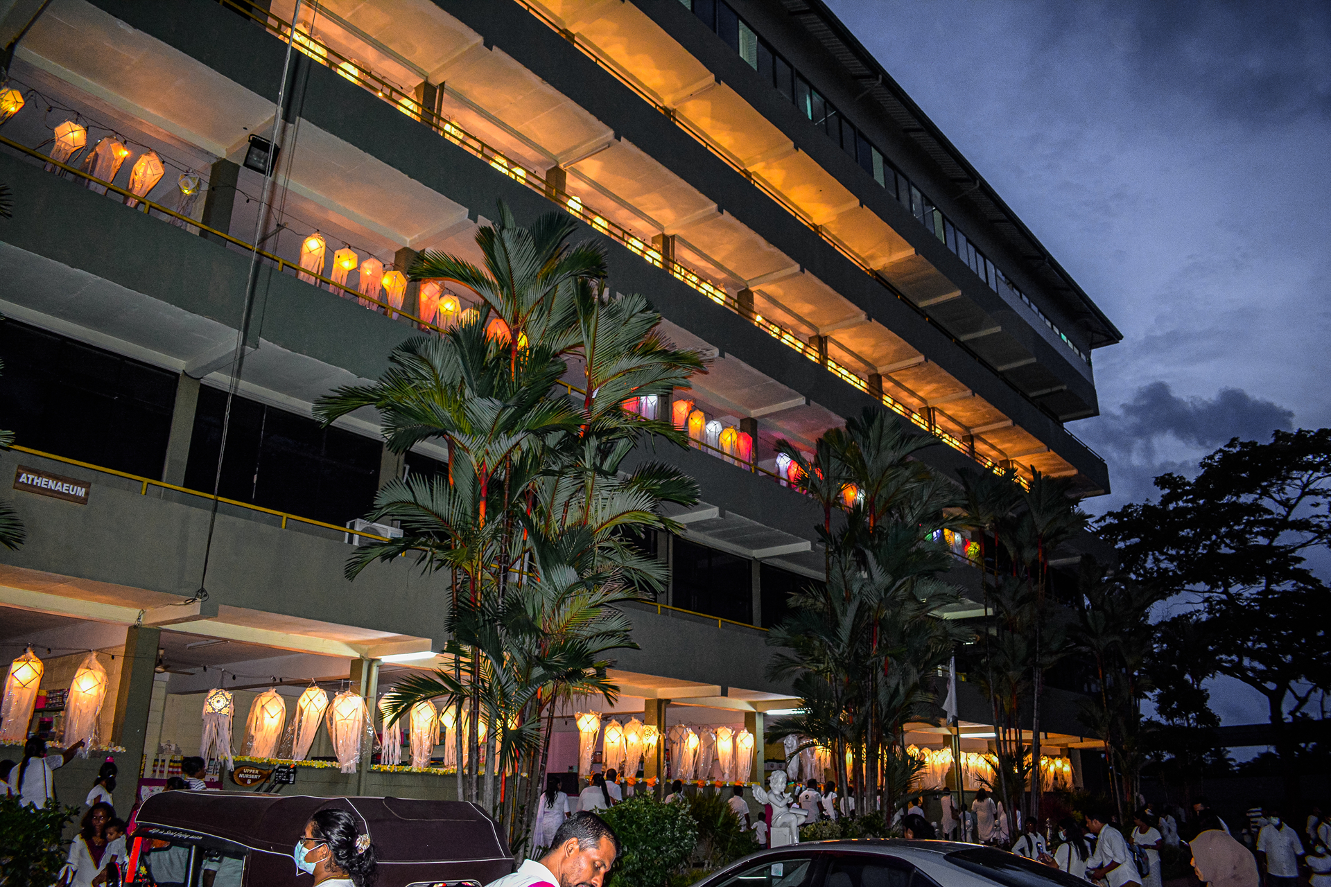 Evening celebration at a multi-story building with lanterns, palm trees, and people in white attire.