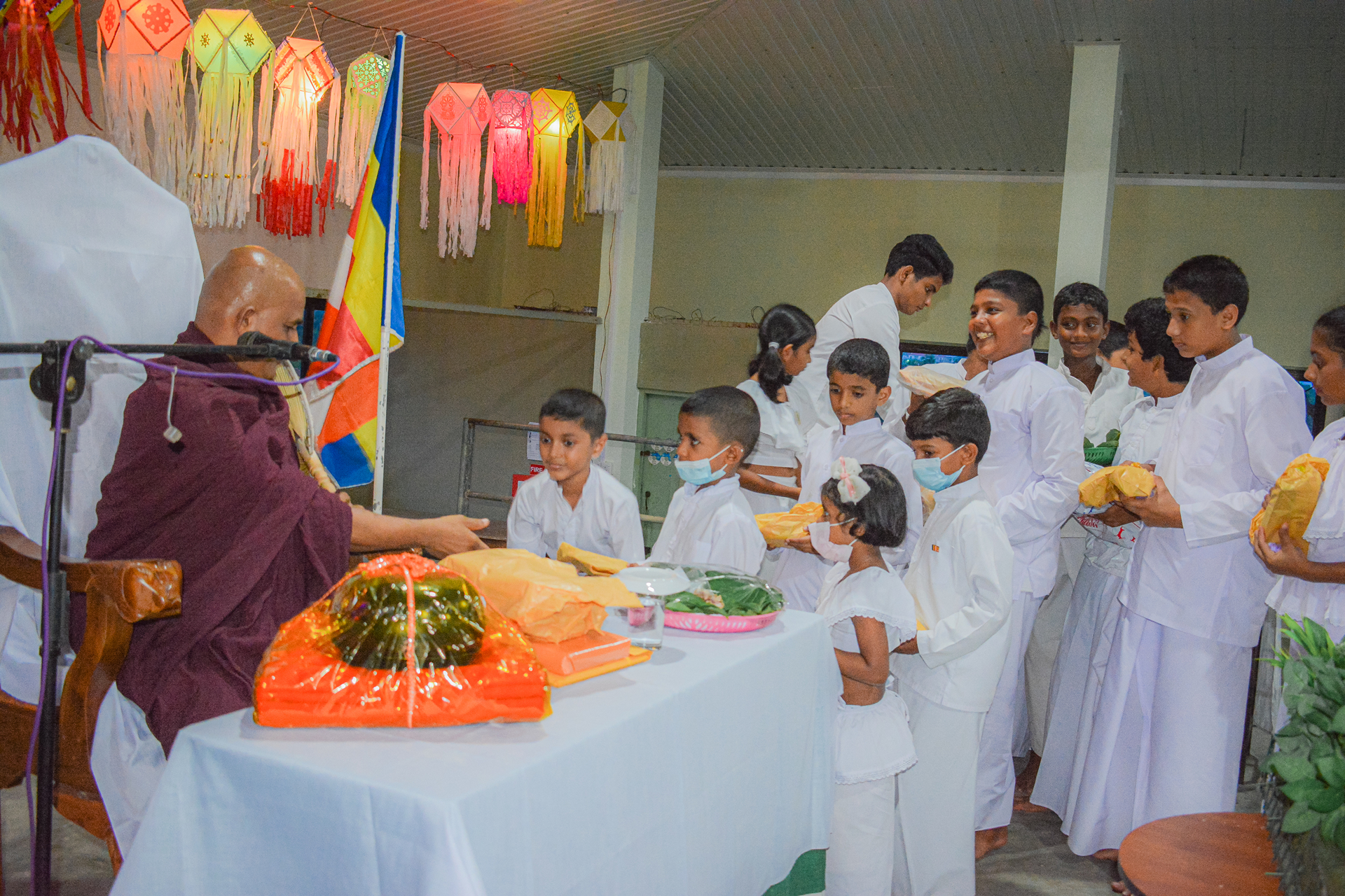 Children offering flowers and gifts to monk during a religious ceremony indoors.