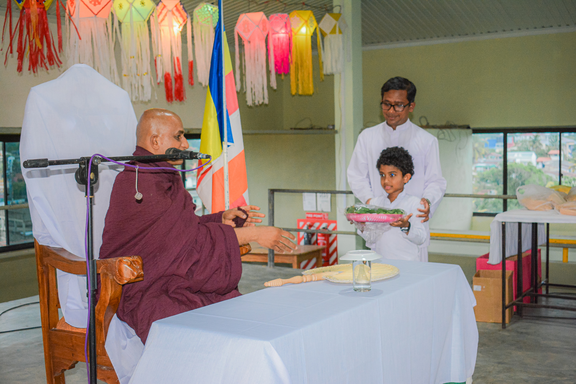 Child offers flowers to monk in maroon robe during colorful ceremonial event.