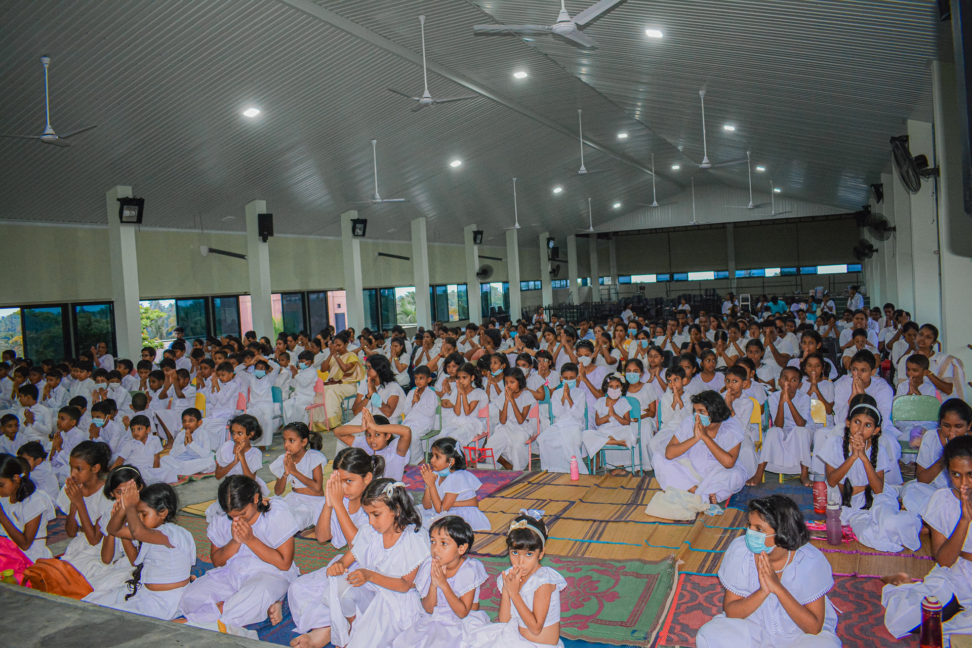 Large group in white praying on colorful mats in a sunlit hall.