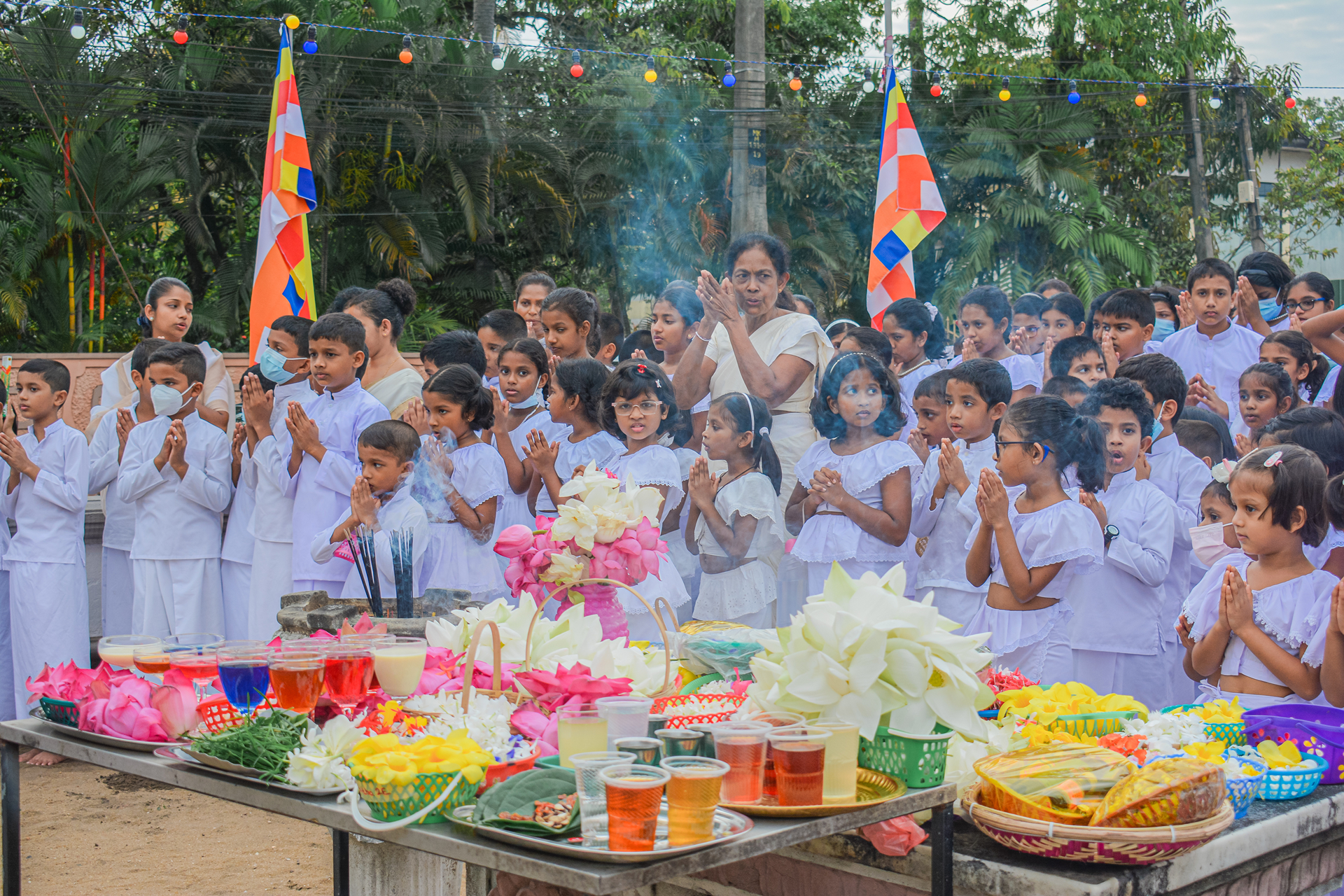 Children in white pray at a Buddhist ceremony with colorful flowers and offerings.