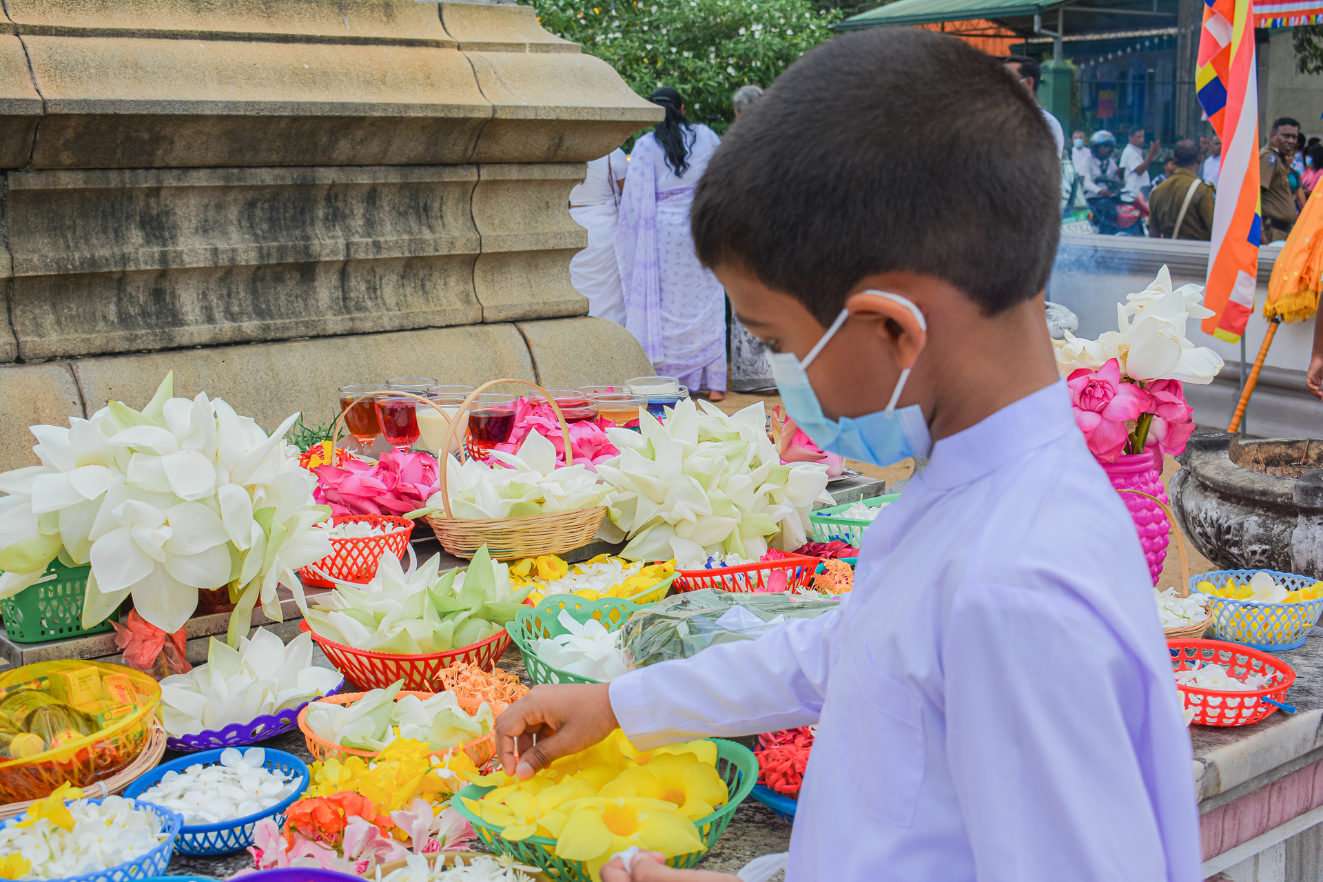 Young boy in white mask arranging colorful offerings at outdoor cultural ceremony.