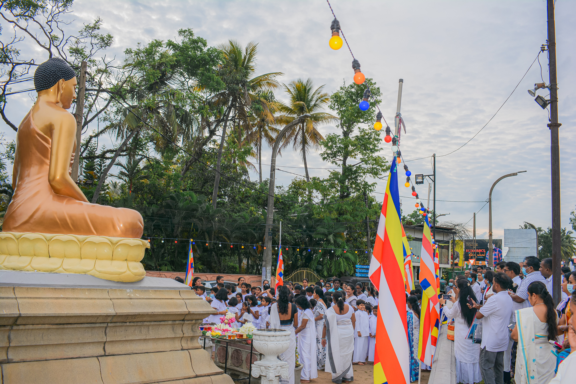 Outdoor Buddhist ceremony with golden Buddha statue, participants in white, and colorful flags.