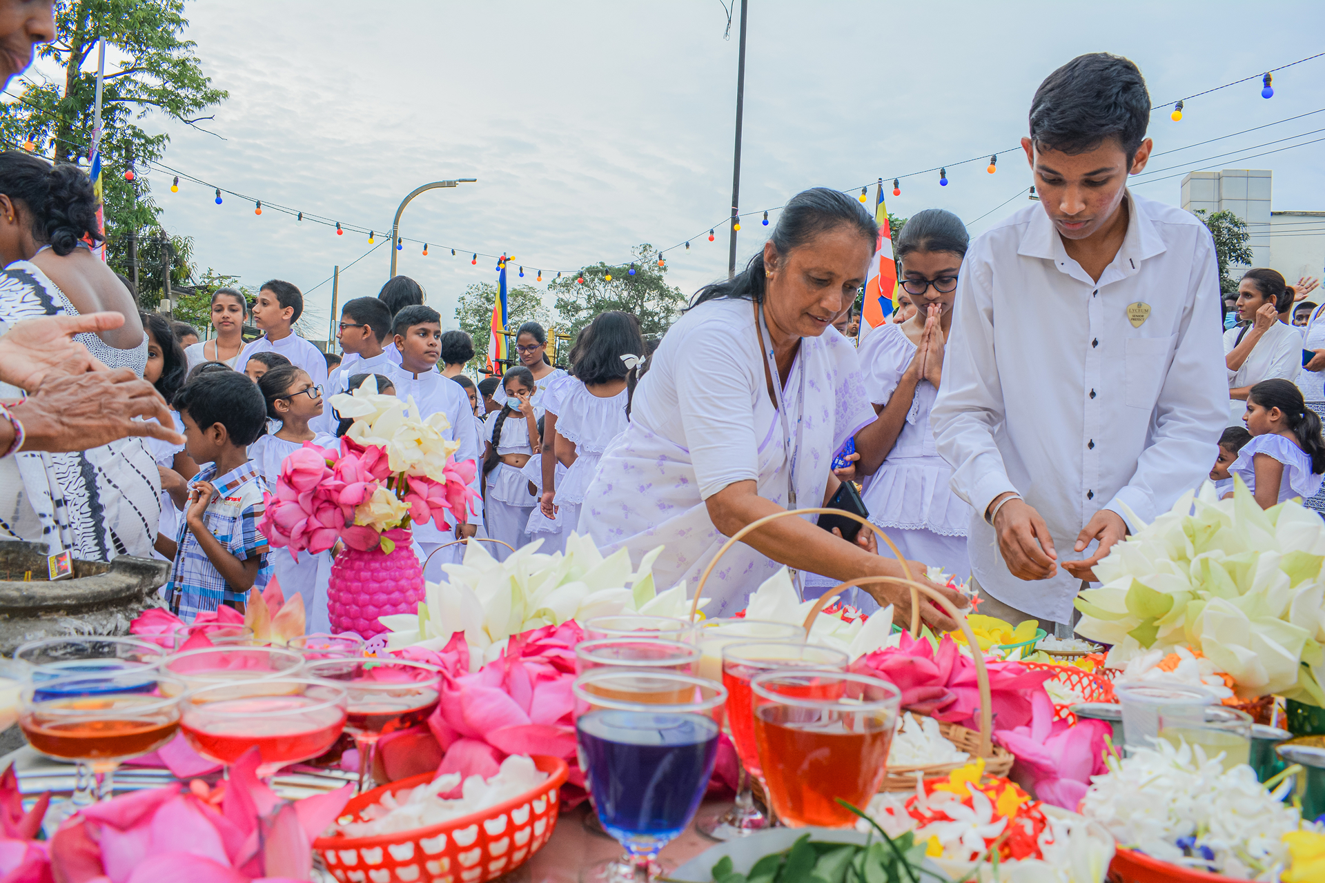 People in white at a cultural event with colorful offerings and flowers.