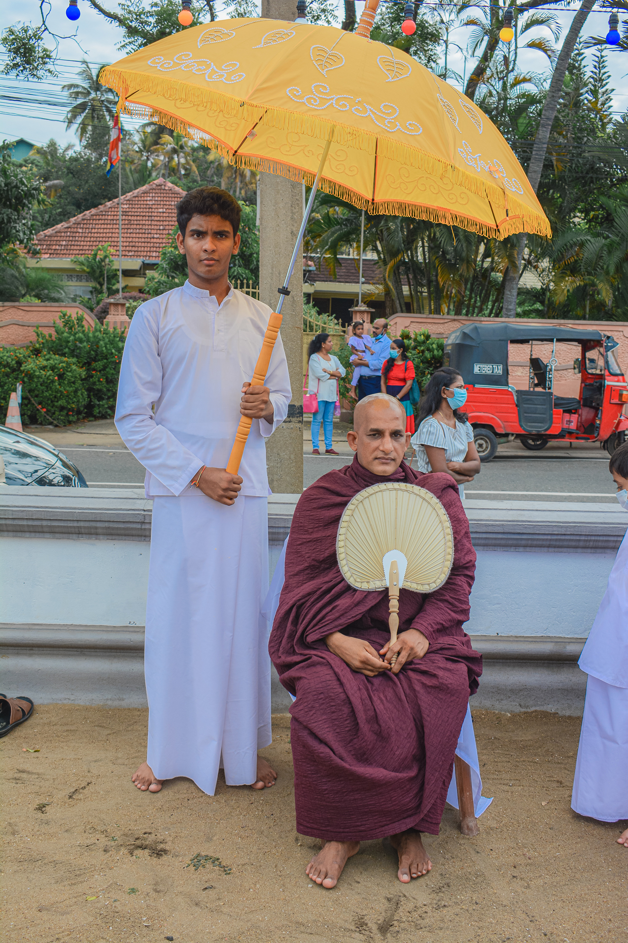 Young man holds umbrella for seated Buddhist monk during ceremony with tuk-tuk in background.