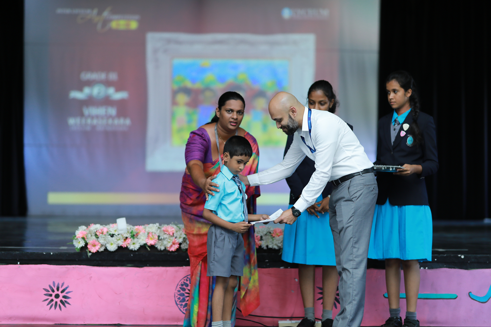 Boy receiving award at school ceremony, supported by a woman, watched by other students.