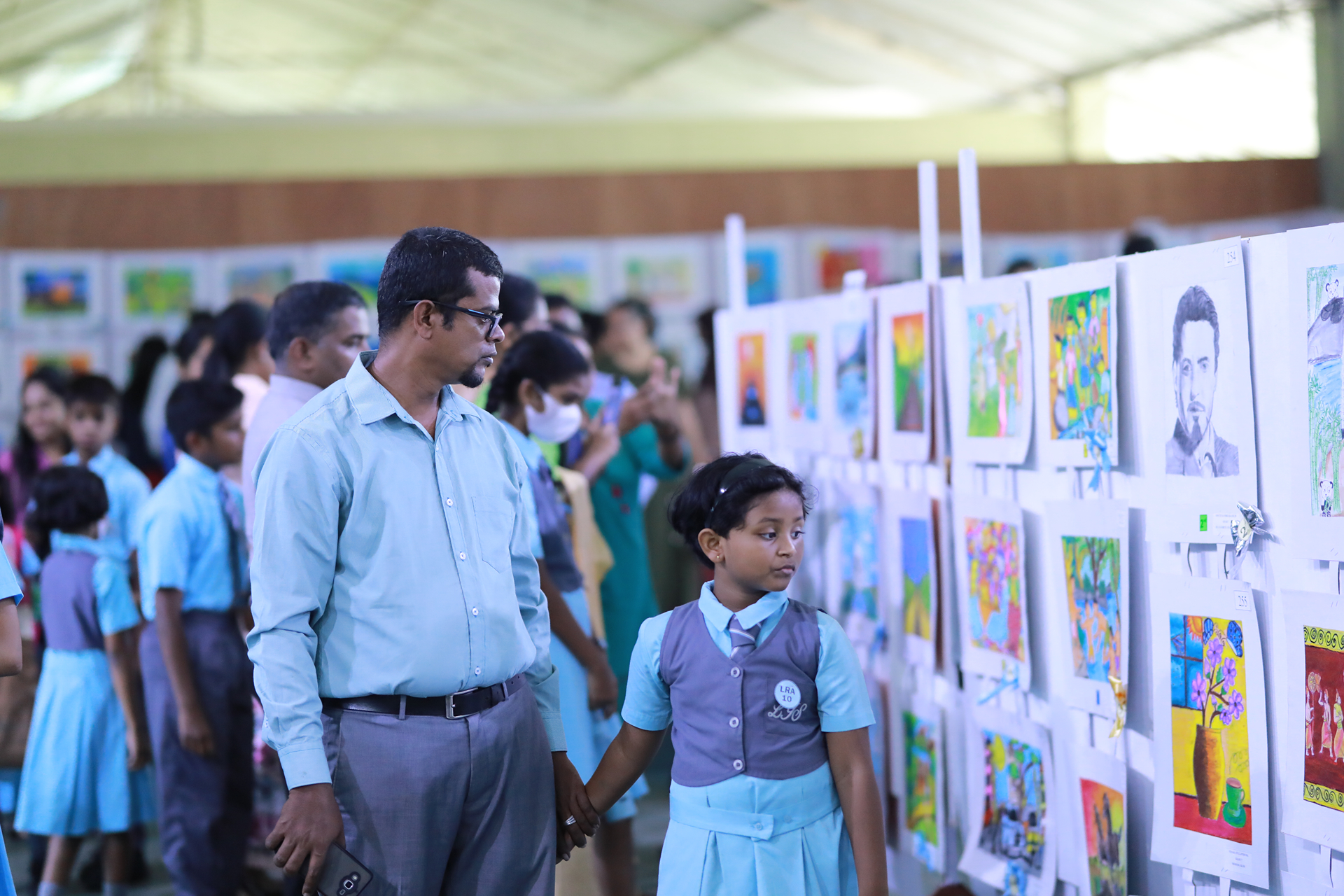 Man and girl admire student artwork at a bustling school art exhibition.