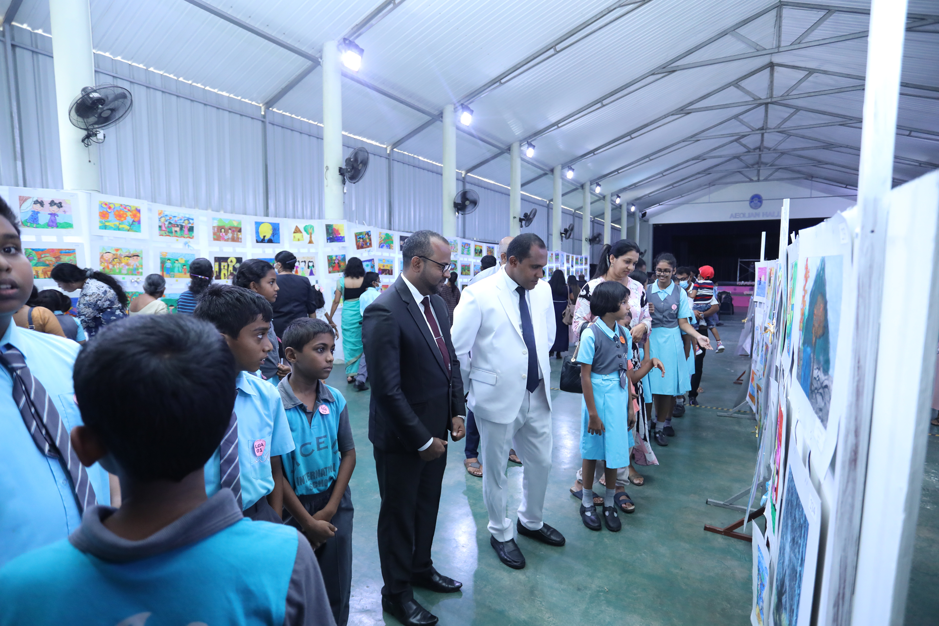 School children and officials viewing art at a busy indoor exhibition.