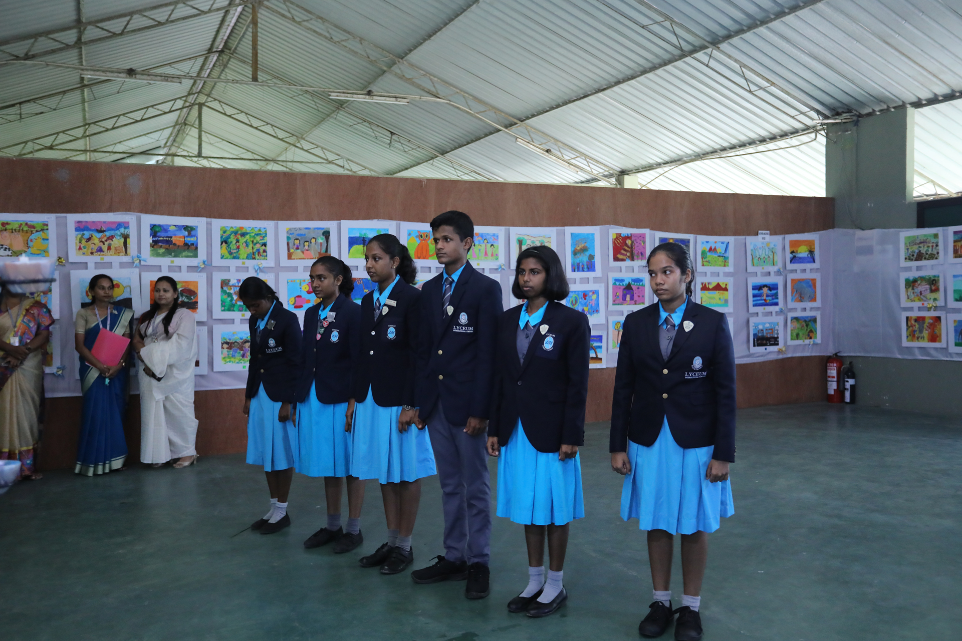 Students at an art exhibition ceremony with colorful artworks on display.