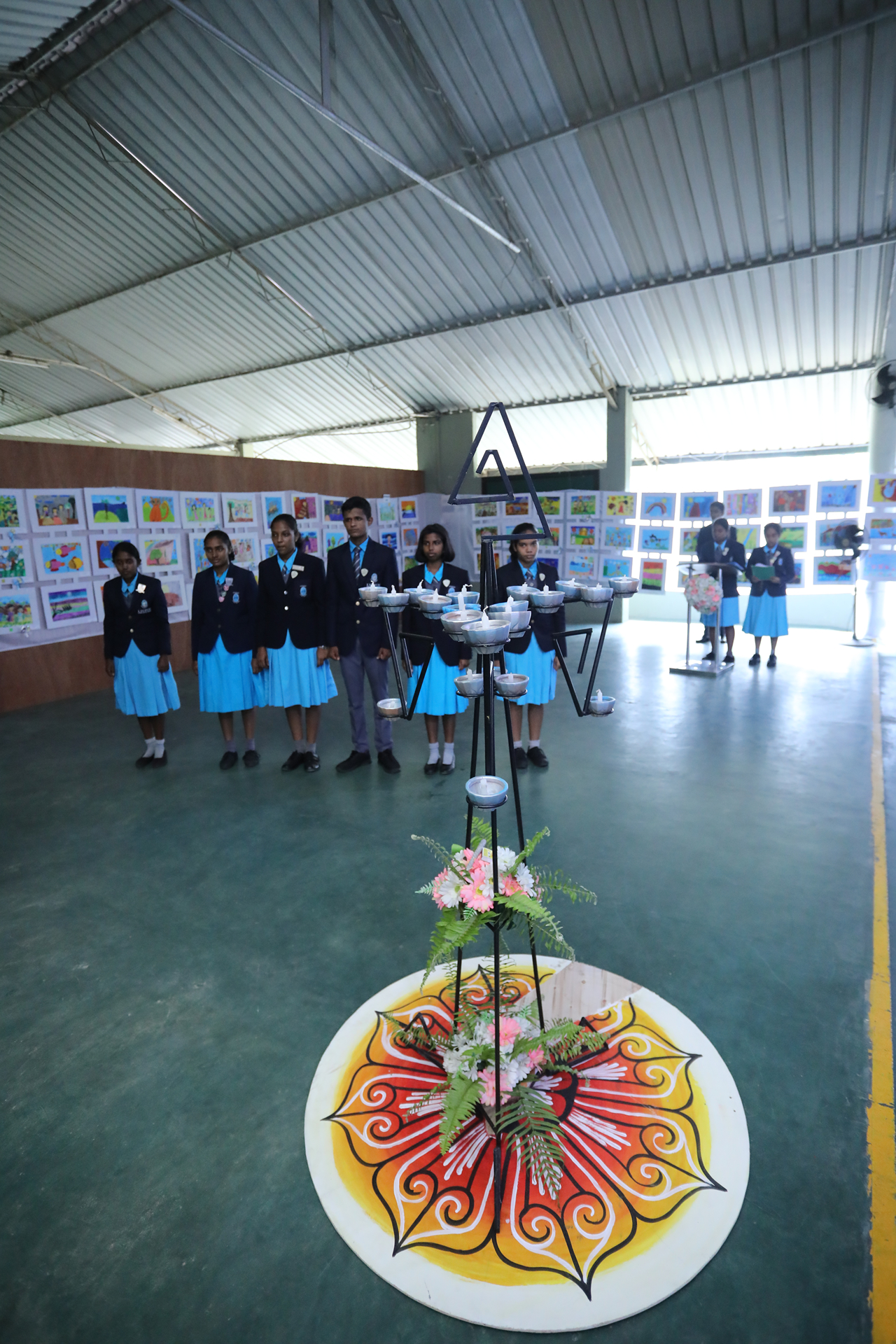 Students in uniforms in a bright school hall with rangoli and art display.