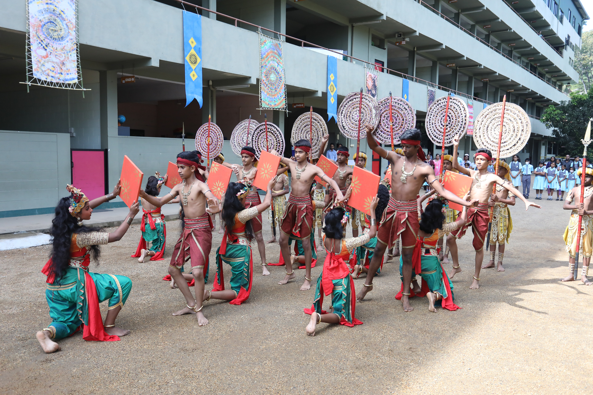 Outdoor cultural dance with colorful costumes and shields in front of a multi-story building.