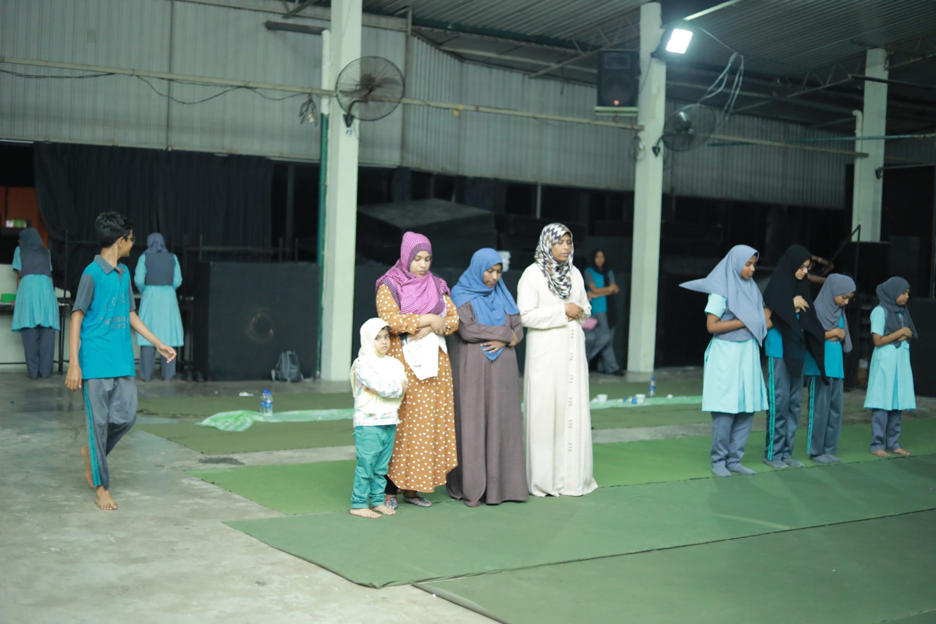 Women and children gather on green mats in a spacious hall for communal prayer.