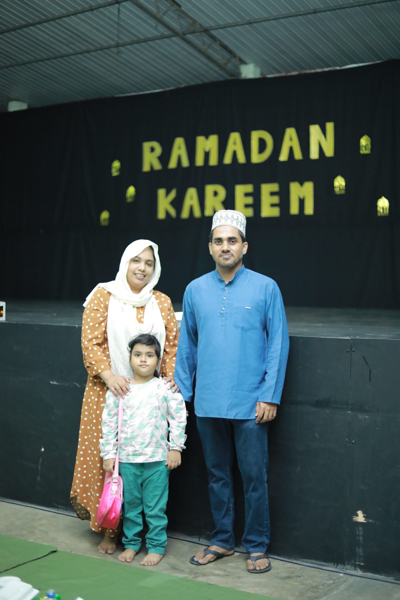 Family of three celebrating Ramadan in traditional attire, standing before a festive Ramadan Kareem backdrop.