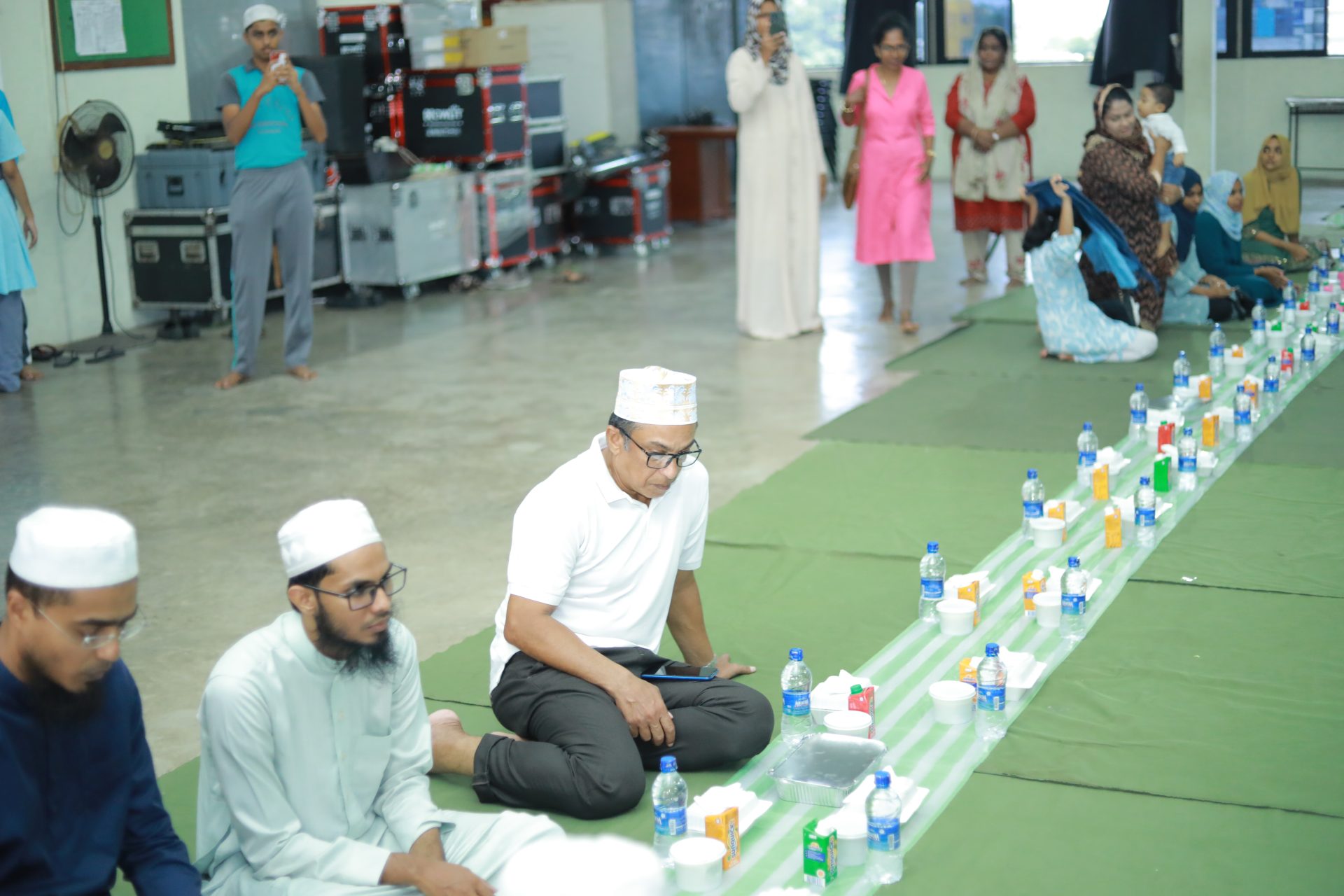 Men in traditional attire seated for Iftar meal in a spacious community hall.