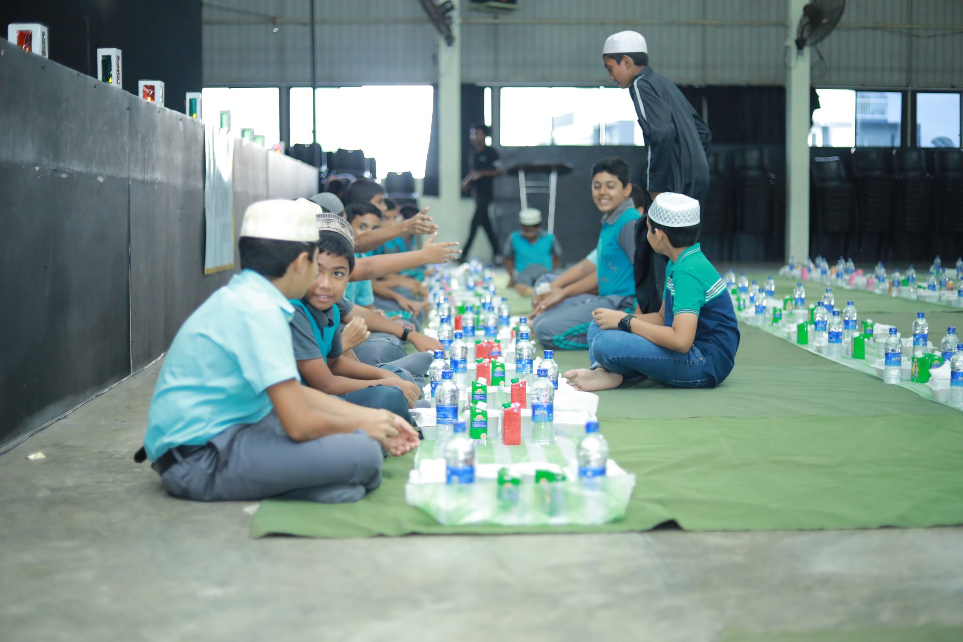 Children in a communal meal, sitting on mats with drinks, in a bright indoor hall.