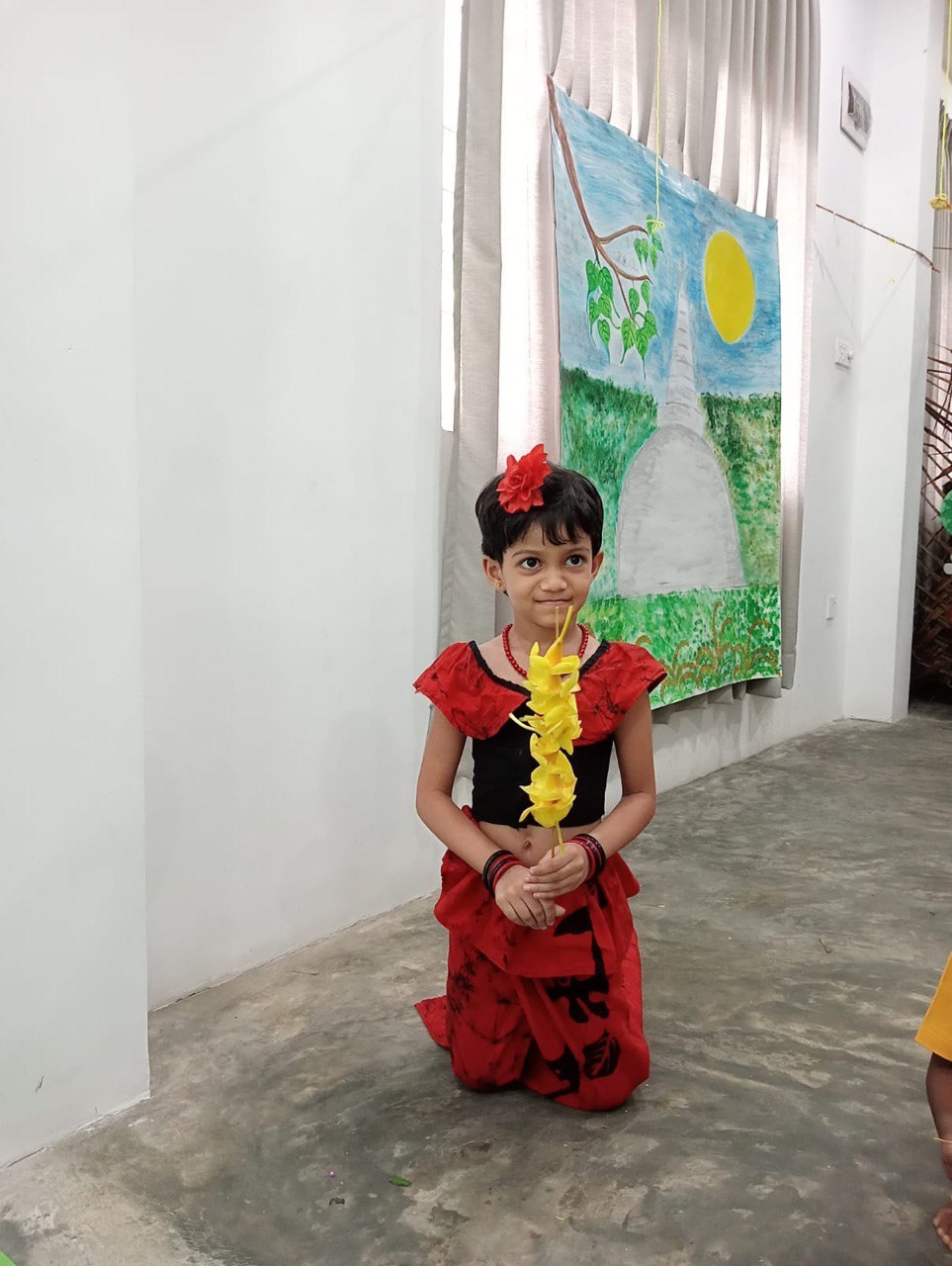 Young child in vibrant traditional attire holding flower garland at cultural event with landscape backdrop.