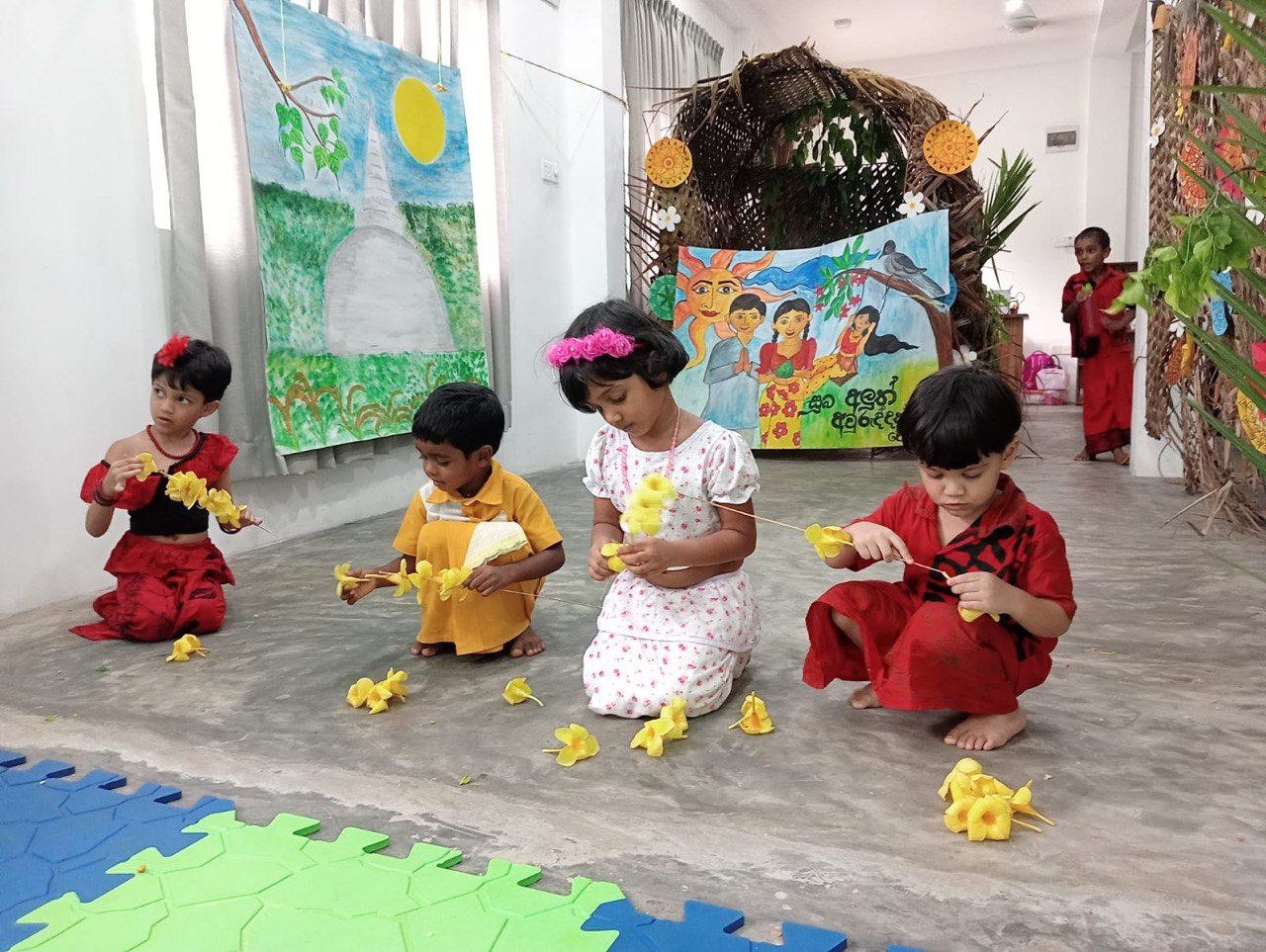 Children in traditional attire arranging flowers in a colorful, art-filled classroom.