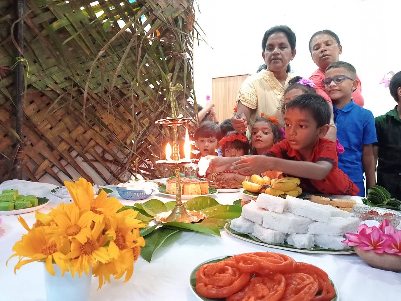 Children and adults celebrate with festive foods and a lit oil lamp on a bamboo backdrop.