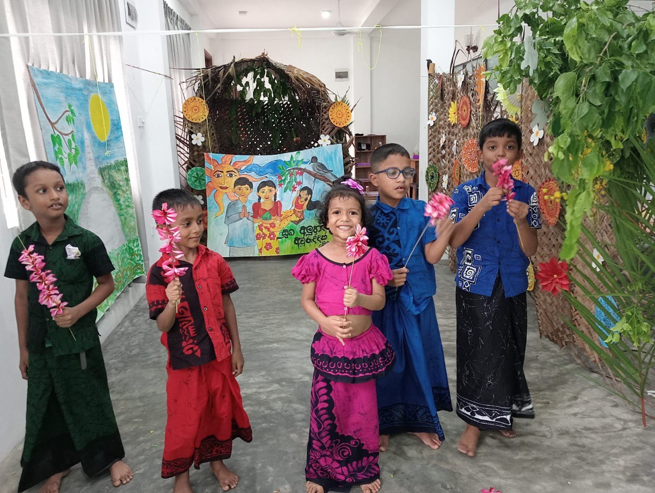 Five children in traditional attire with floral garlands, festive indoor setting.