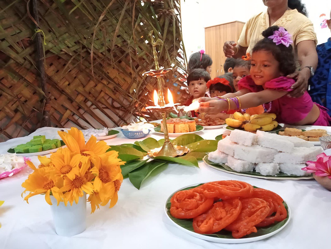 Traditional celebration with children, colorful attire, lit oil lamp, and assorted foods on a decorated table.