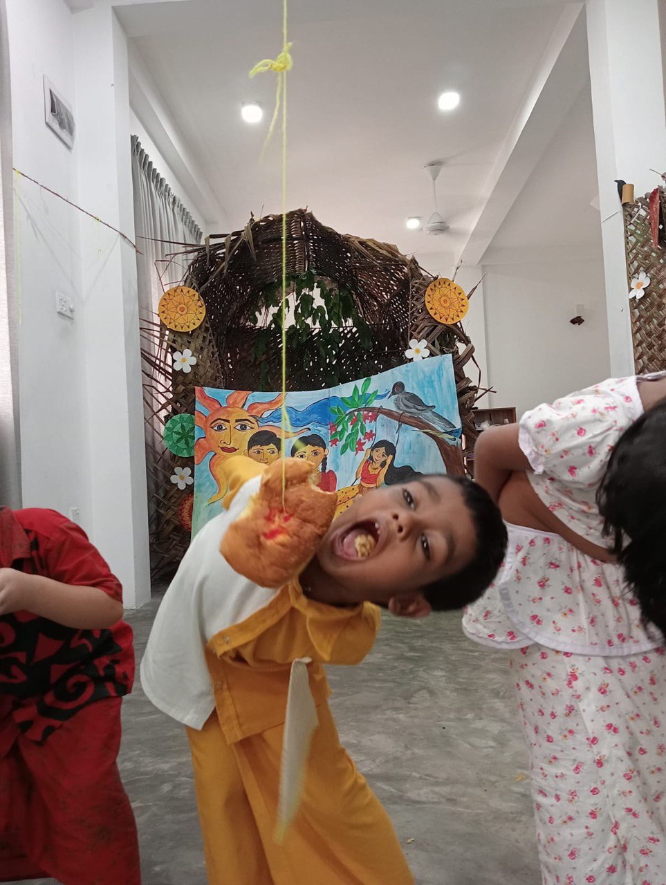 Children enjoying a festive game, trying to bite hanging treats in a colorful, decorated room.