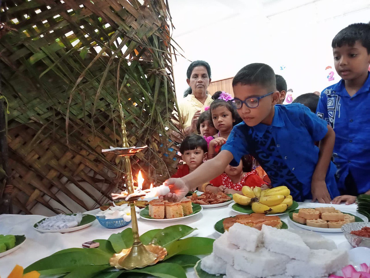 Children celebrating a cultural ceremony with traditional foods and lighting a brass oil lamp.