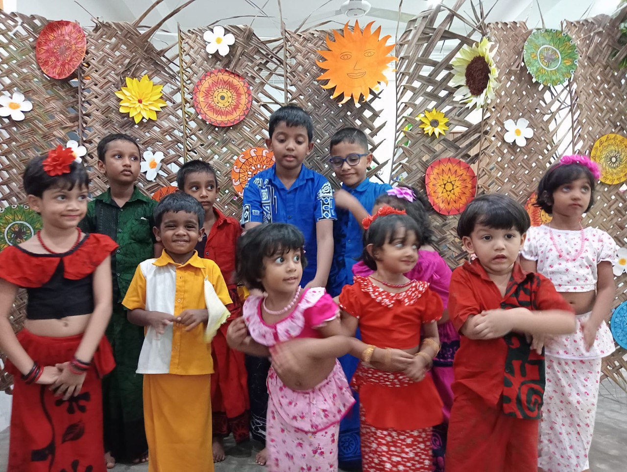 Eleven children in traditional festive attire posing before a colorful, flower-adorned backdrop.